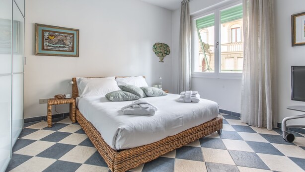 A neatly arranged bedroom with a checkered floor, a wicker bed, a bedside table, a wall-mounted TV, and a window with curtains. - Venezia - Lido Erizzo 15 (Venice)