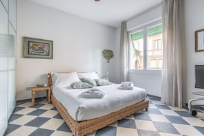 A neatly arranged bedroom with a checkered floor, a wicker bed, a bedside table, a wall-mounted TV, and a window with curtains. - Venezia - Lido Erizzo 15 (Venice)