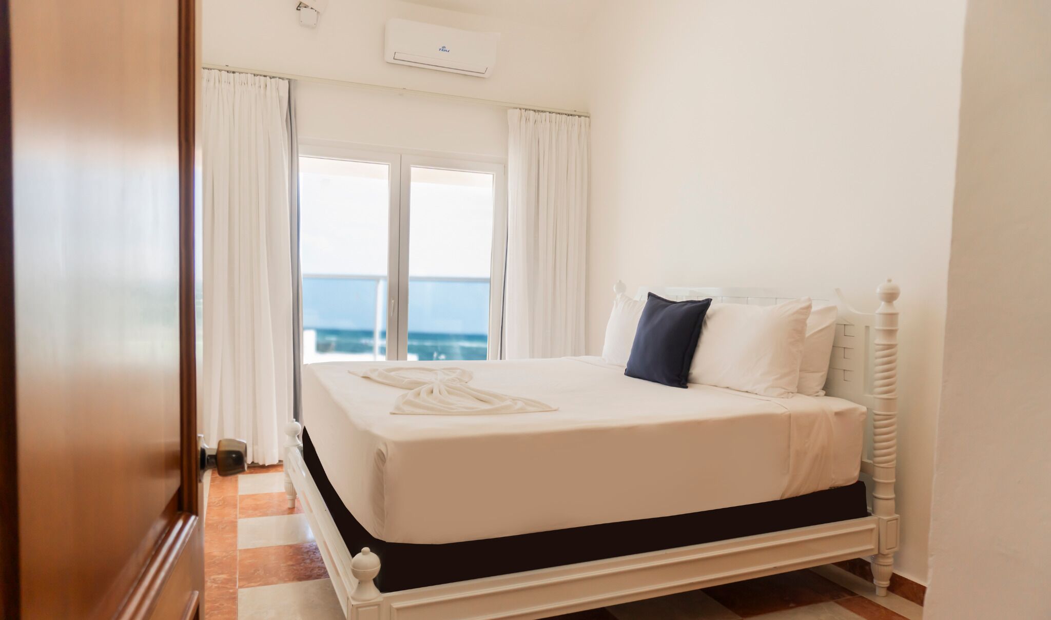 A hotel room with a bed, white curtains, a view of the sea, and an air conditioning unit.