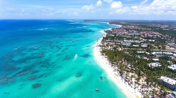 Luchtbeeld van een kustgebied met een strand, helder turquoise water en een dichtbebouwd woongebied met gebouwen en groen.