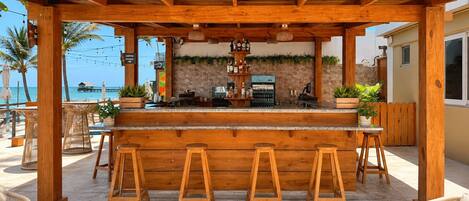 A wooden bar with stools, a stone wall, and a view of the beach.