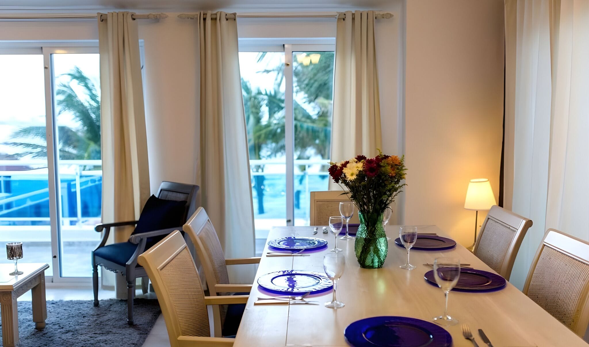 A dining area with a table set for four, a vase with flowers, and a view of a pool and palm trees through large windows.