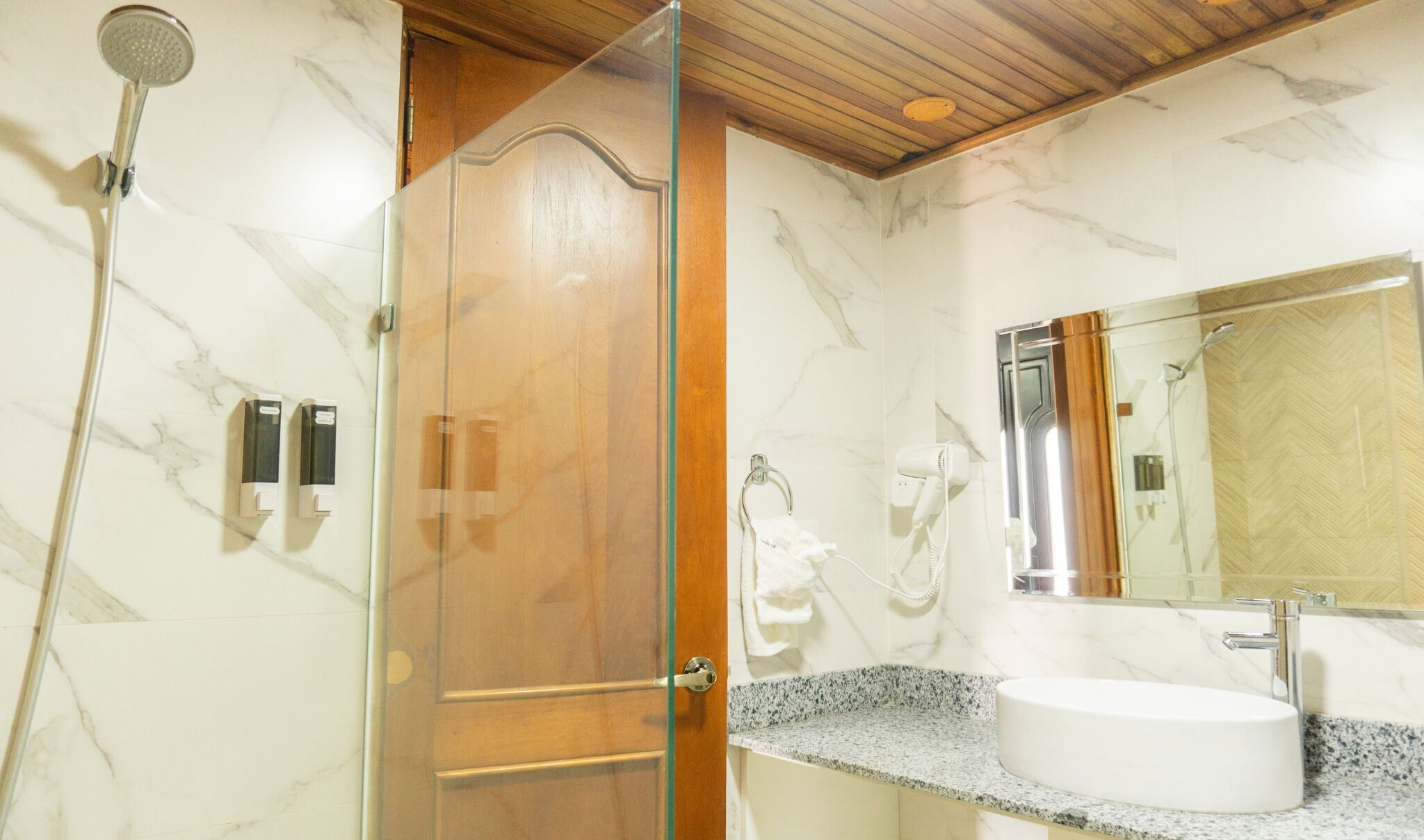 A bathroom with a glass shower enclosure, a wooden door, a marble wall, a sink with a granite countertop, and a mirror with a wooden frame.