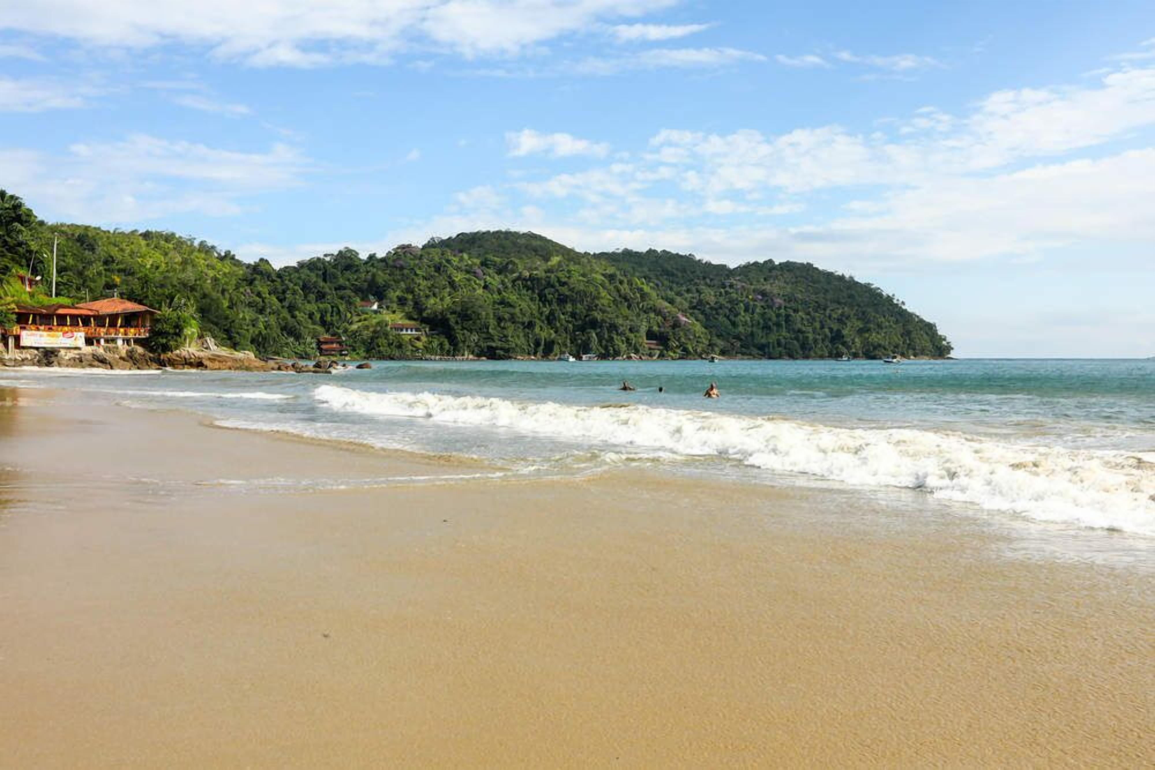 Ein Strand mit einem Gebäude links, einer bewaldeten Hügelkette im Hintergrund und ruhigen Wellen.