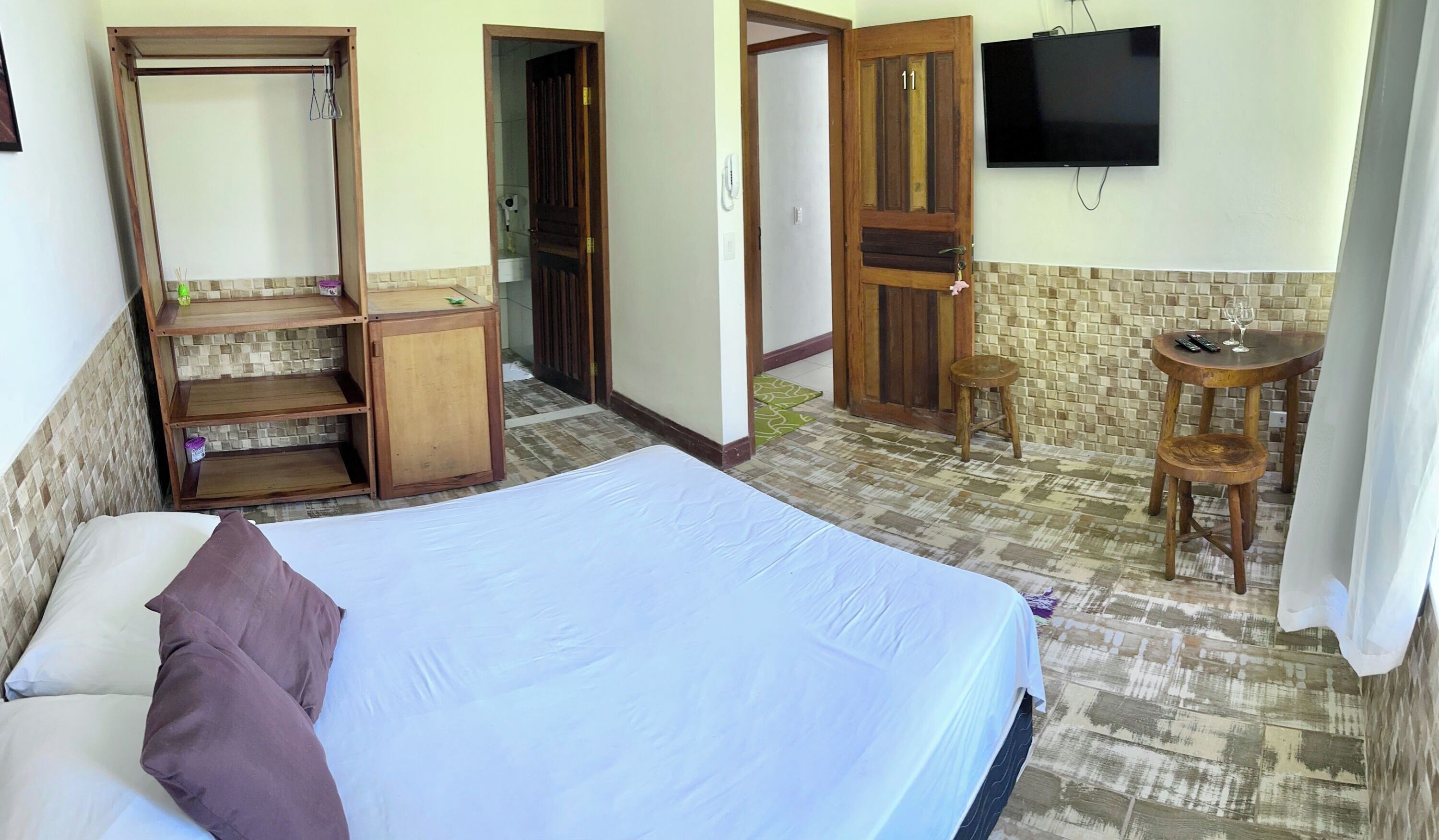 Quarto com uma cama, um guarda-roupa de madeira, uma mesinha com uma cadeira, uma TV e uma porta com o número 11.