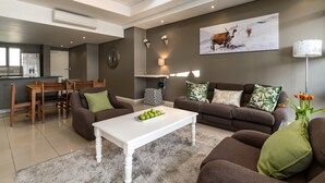 A modern living room with a brown sofa, white coffee table, and a dining area with wooden furniture. - Flatrock 909 (Cape Town)