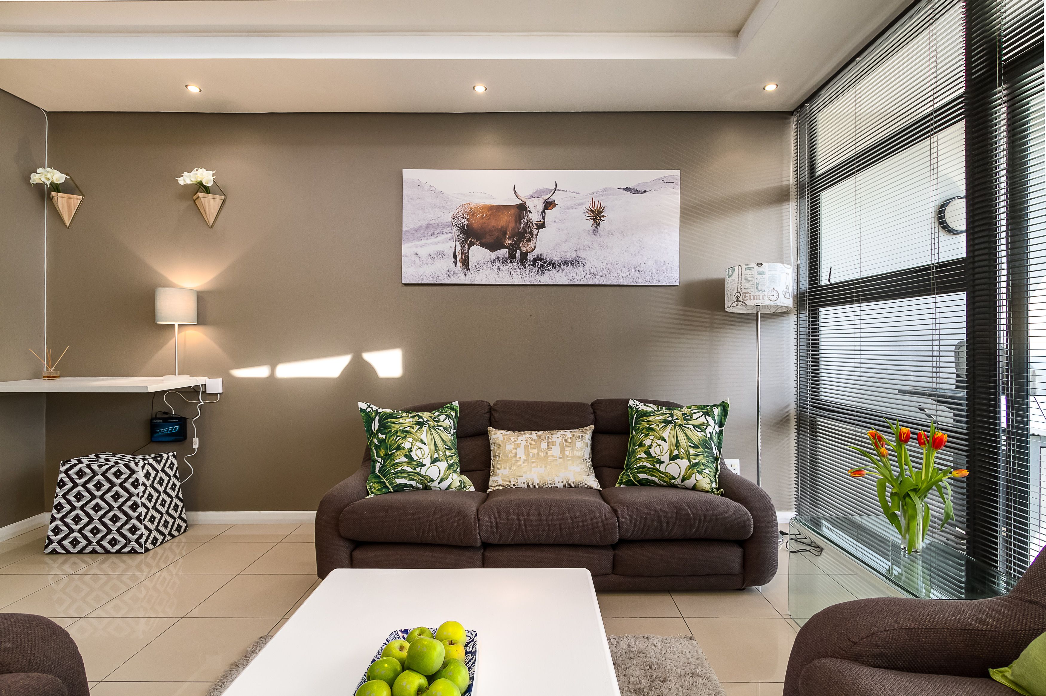 A modern living room with a brown sofa, a coffee table with green apples, a wall-mounted picture of a bull, and a window with orange tulips.