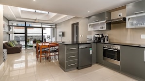 A modern kitchen with a built-in dishwasher, oven, and coffee maker. There is a dining area with a table and chairs. - Flatrock 909 (Cape Town)