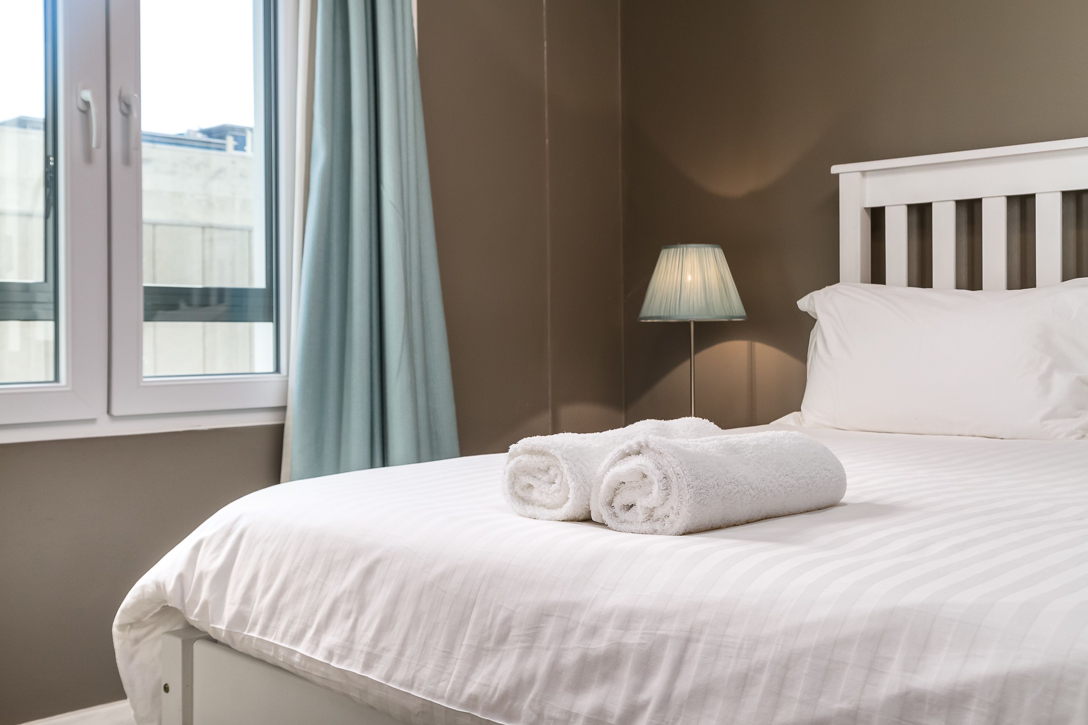 A neatly made bed with white linens and rolled towels, a bedside lamp, and a window with curtains.
