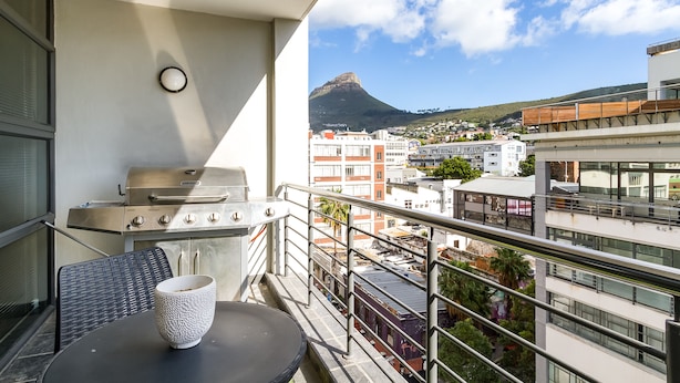A balcony with a grill, a table with a bowl, and a view of a mountain and buildings. - Flatrock 909 (Cape Town)