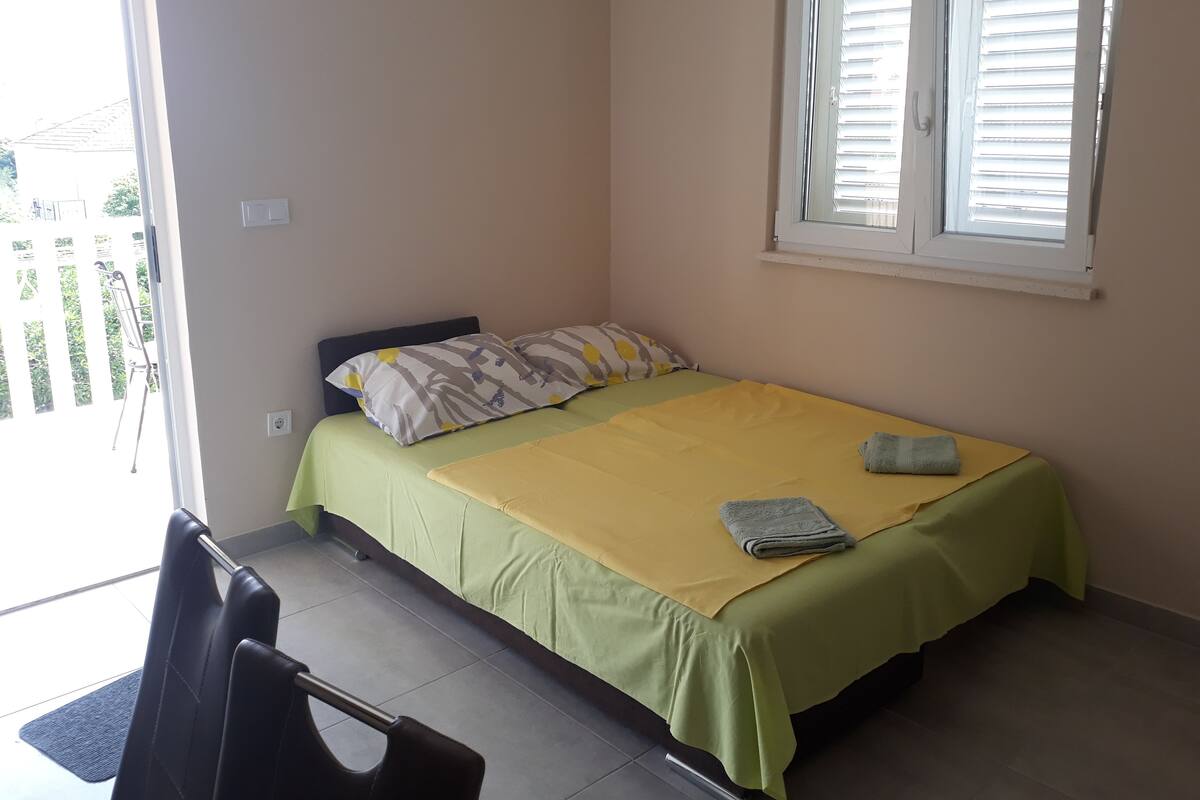 A single bed with a green and yellow bedspread, two pillows, a folded towel, and a window with blinds.