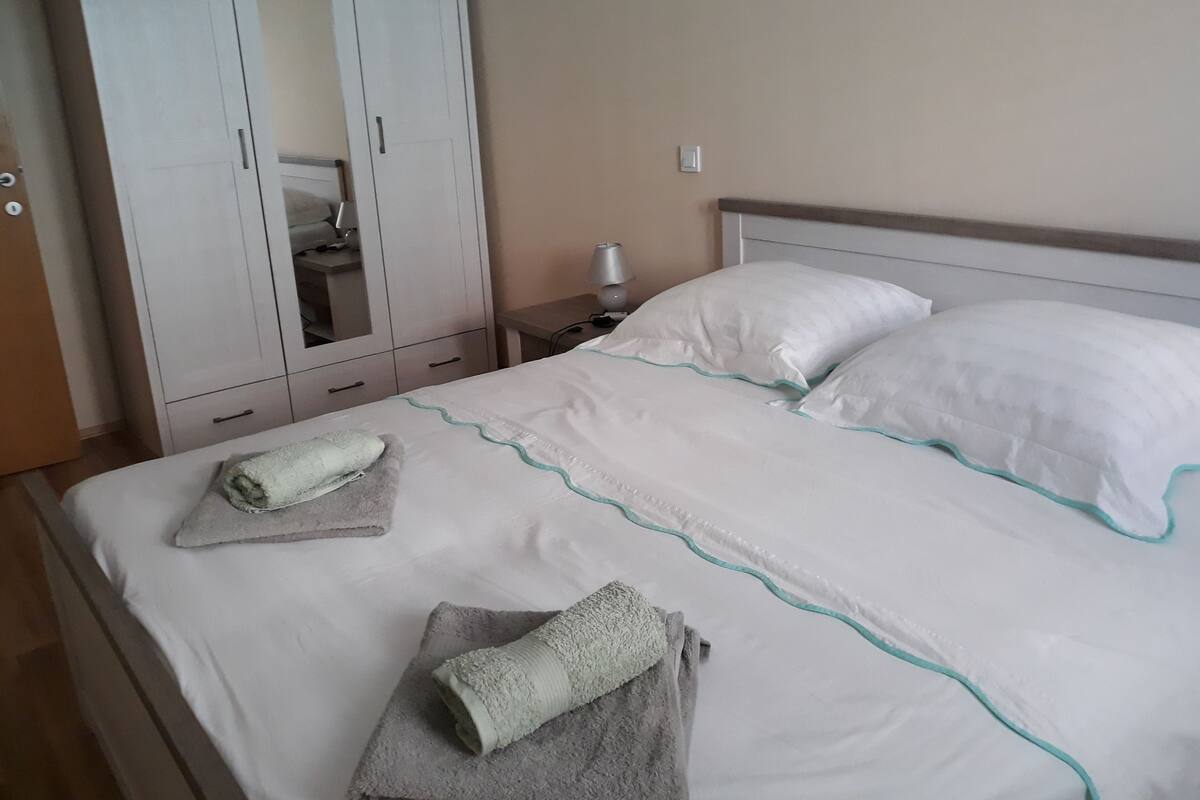 A neatly made bed with white bedding and teal trim, a grey towel on the bed, and a white wardrobe with a mirror.