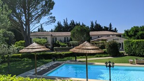A swimming pool with a clear blue water, surrounded by well-maintained greenery and a large tree. - Le Pradet Hotel (Vacqueyras)