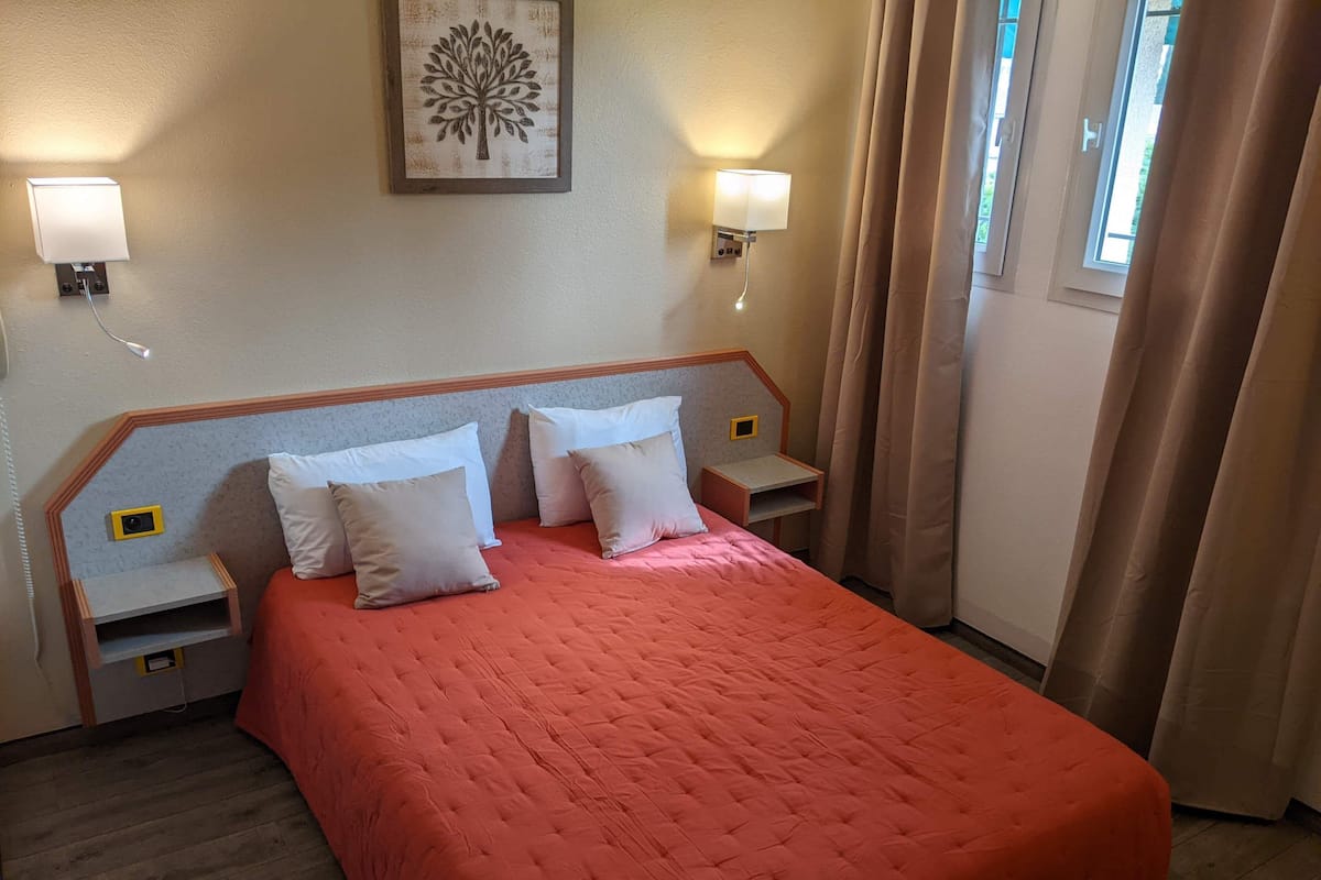 A hotel room with a large bed, two bedside tables, a wall-mounted lamp, and a window with curtains.