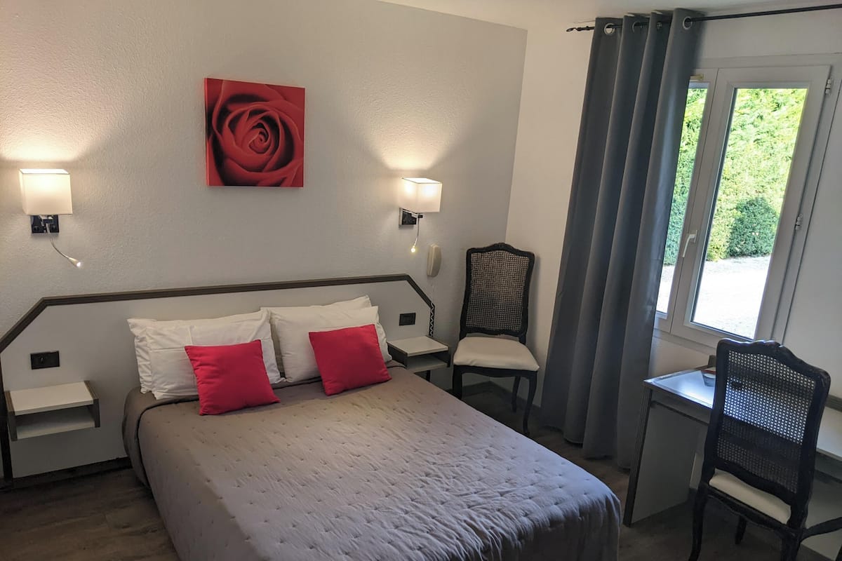 A bedroom with a bed, two bedside tables, a chair, a desk, a window with curtains, and a wall-mounted artwork.