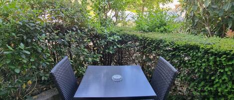 A black metal table with a clear glass center, surrounded by green foliage and a hedge.