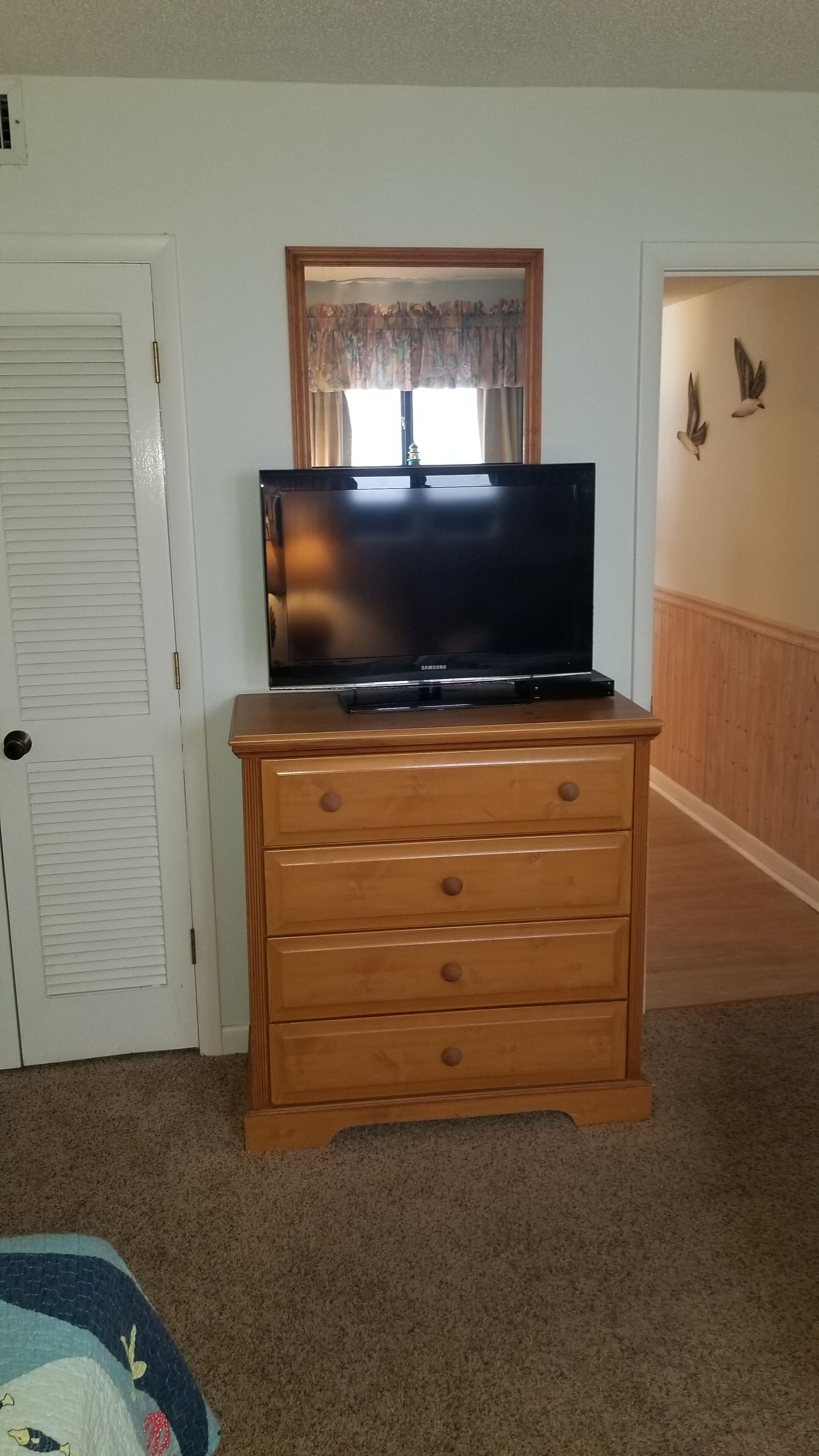 Quarto com cômoda de madeira, televisão em cima, porta com venezianas, janela com cortina e parede decorada com dois pássaros.