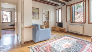 A living room with a blue sofa, a large patterned rug, wooden beams on the ceiling, and a window with a view of a tiled roof. - Venezia - Castello 1542 (Venice)