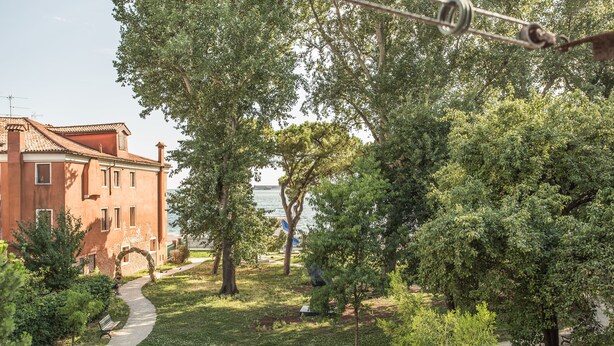 A pathway leading to a building with a red facade, surrounded by lush greenery and trees. - Venezia - Castello 1542 (Venice)
