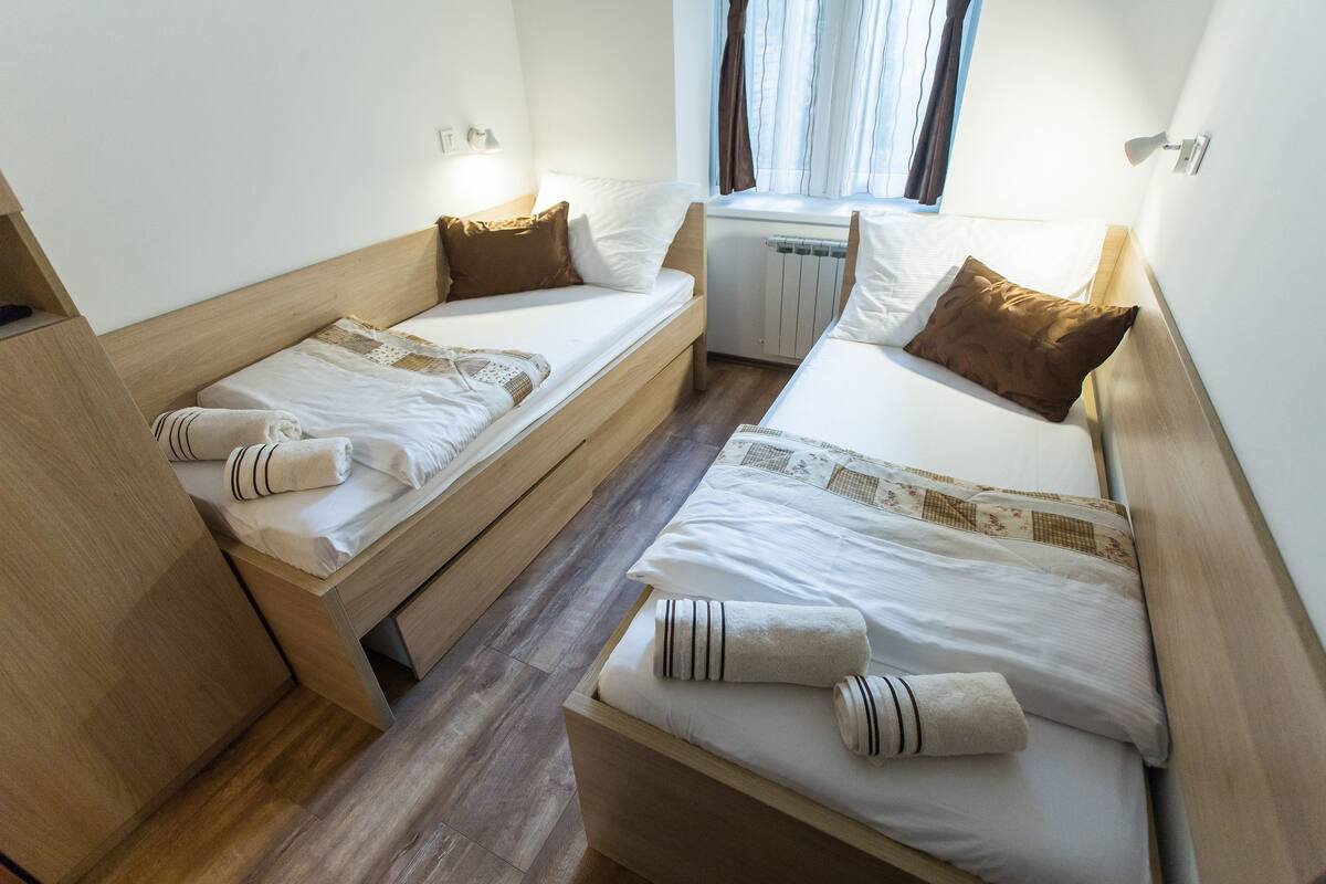 A compact hotel room with two single beds, wooden headboards, and a window with curtains.