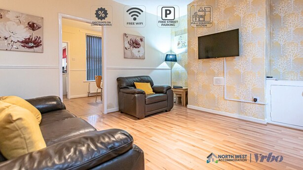 Ein modernes Wohnzimmer mit einer Couch, einem Couchtisch und einem an der Wand montierten Fernseher. An den Wänden befinden sich florale Kunstwerke und eine gemusterte Tapete. Der Raum hat einen Holzboden und eine Tür, die in einen anderen Bereich führt.