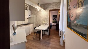 Une cuisine moderne avec un mur de pierres apparentes derrière le plan de travail, un coin repas avec une table et des chaises blanches, et une œuvre d’art encadrée accrochée au mur.