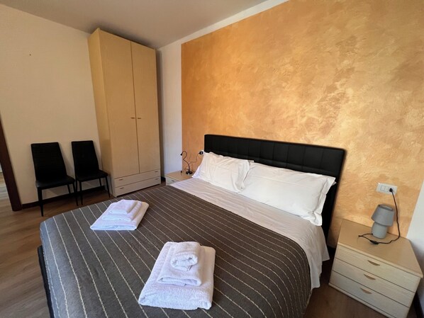 Habitación de hotel con una cama, dos sillas, un armario y una mesita de noche con lámpara.