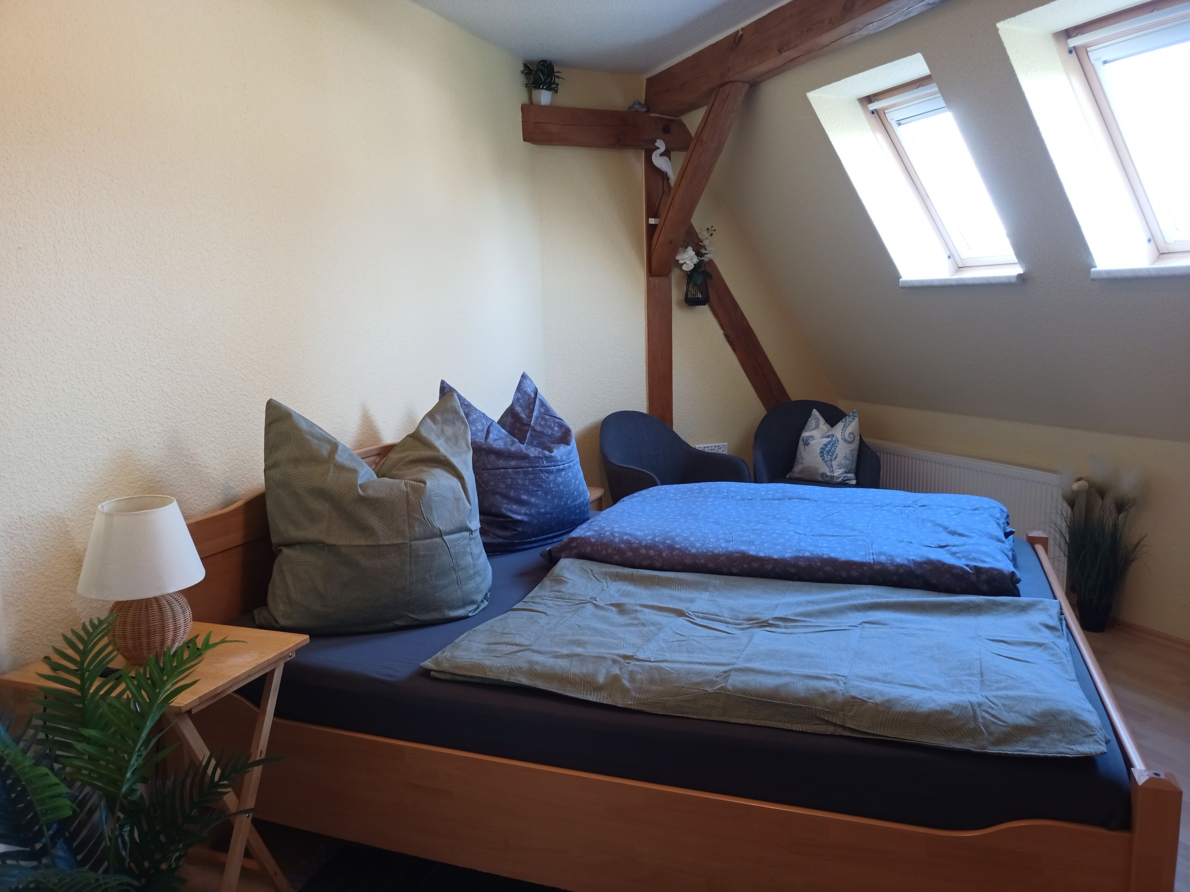 A bedroom with a wooden bed, multiple pillows, a bedside lamp, a window, and a plant.