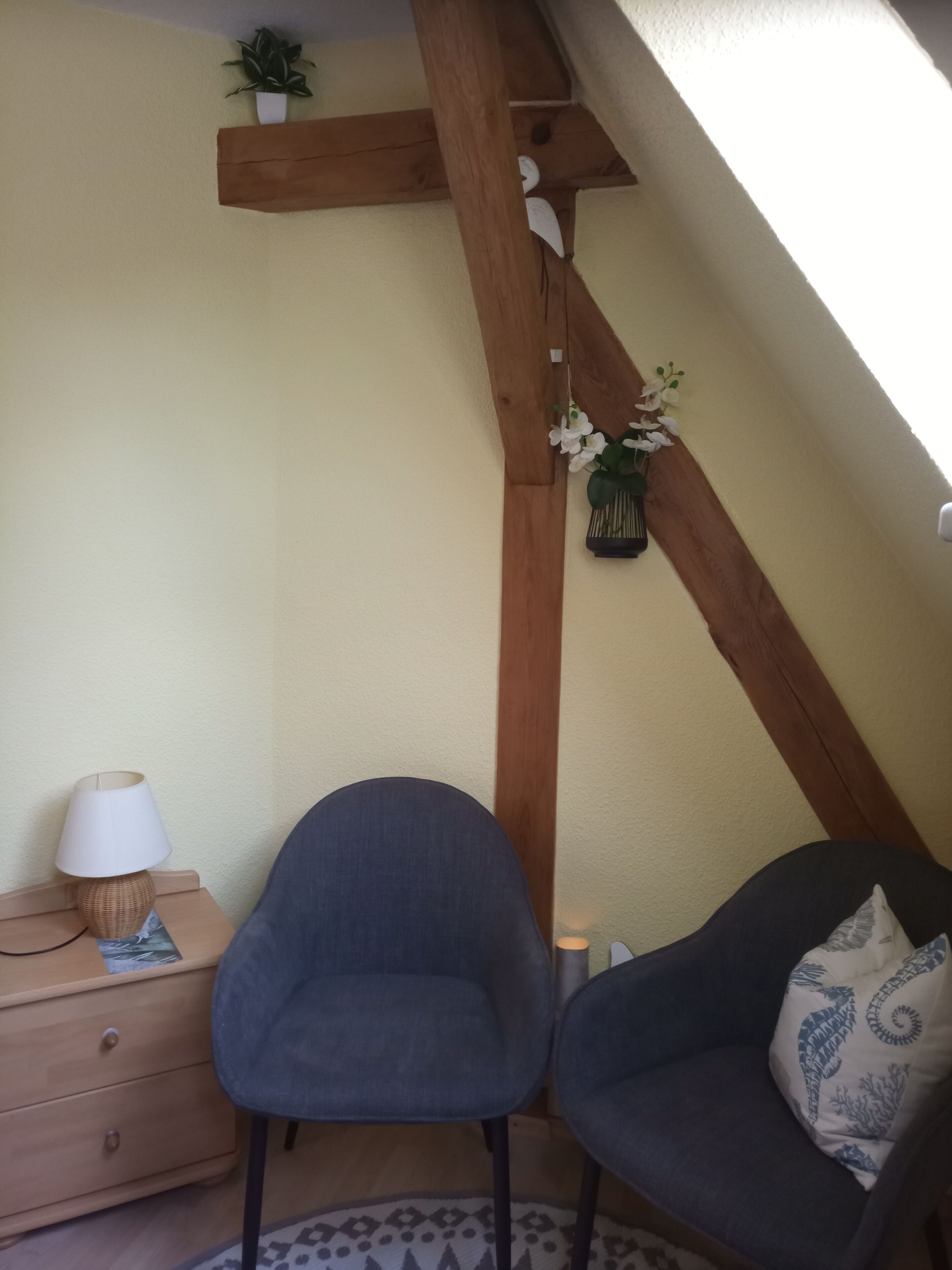 Una habitación con techo de vigas de madera, una silla, una mesita con lámpara y una planta decorativa.