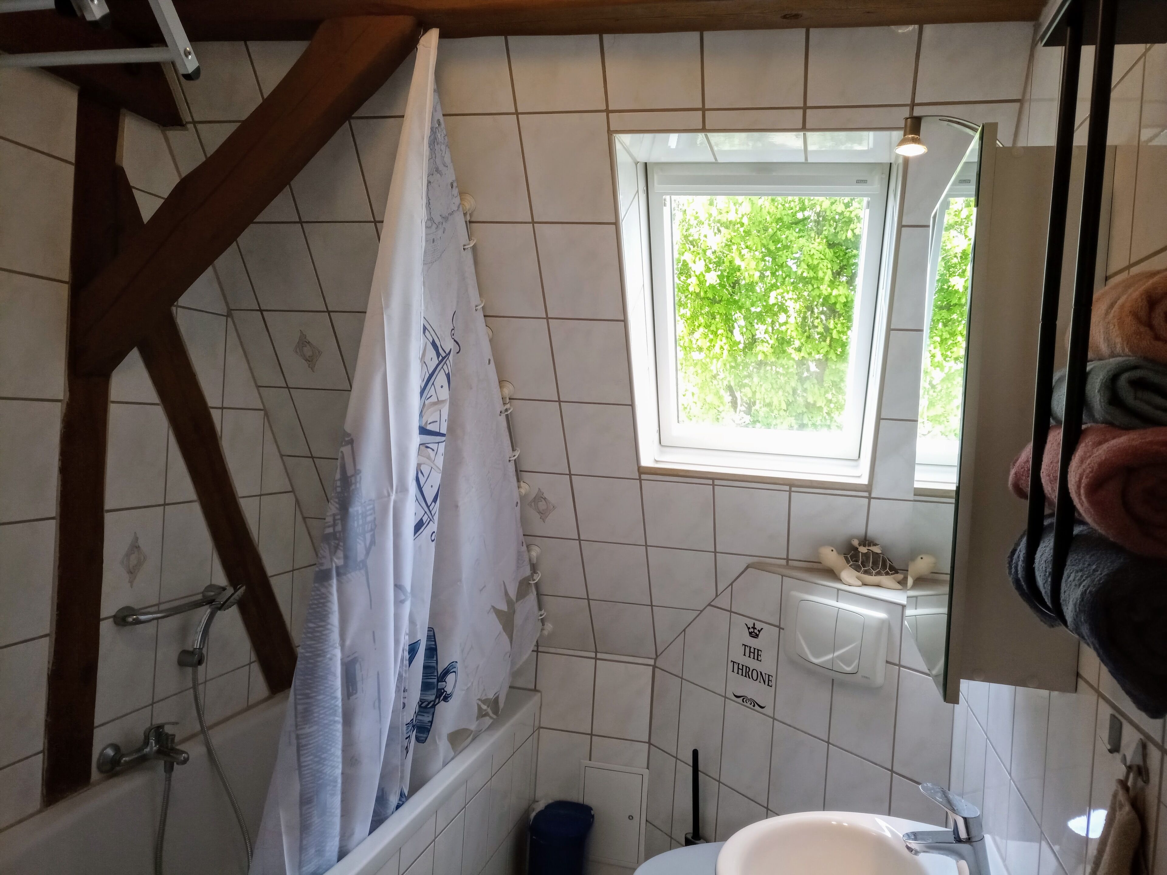 Un baño con ducha, una ventana con vistas a la vegetación, un cartel que dice "THE THRONE" y un perchero con toallas.