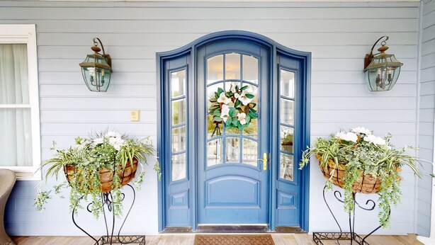 Une porte bleue avec un panneau de verre, flanquée de deux lanternes fixées au mur et de deux plantes en pot.