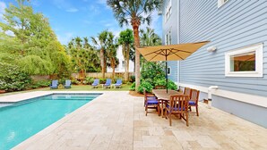 Un jardin avec une piscine, des chaises longues, une table en bois avec des chaises et un grand parasol.