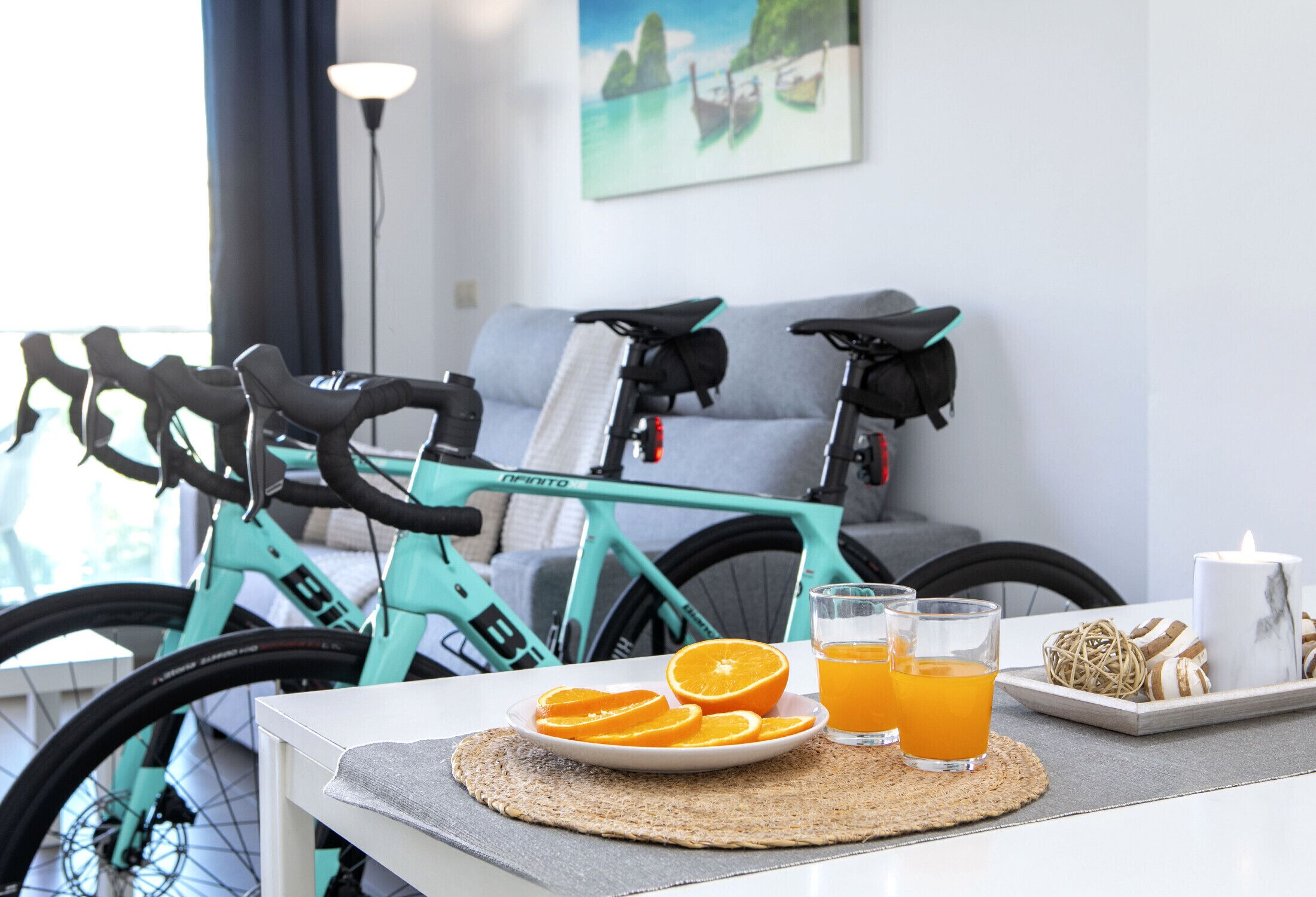 Une pièce dans laquelle on aperçoit un vélo de couleur turquoise, une assiette de tranches d’orange, deux verres de jus d’orange, une bougie et un objet décoratif posé sur une table.