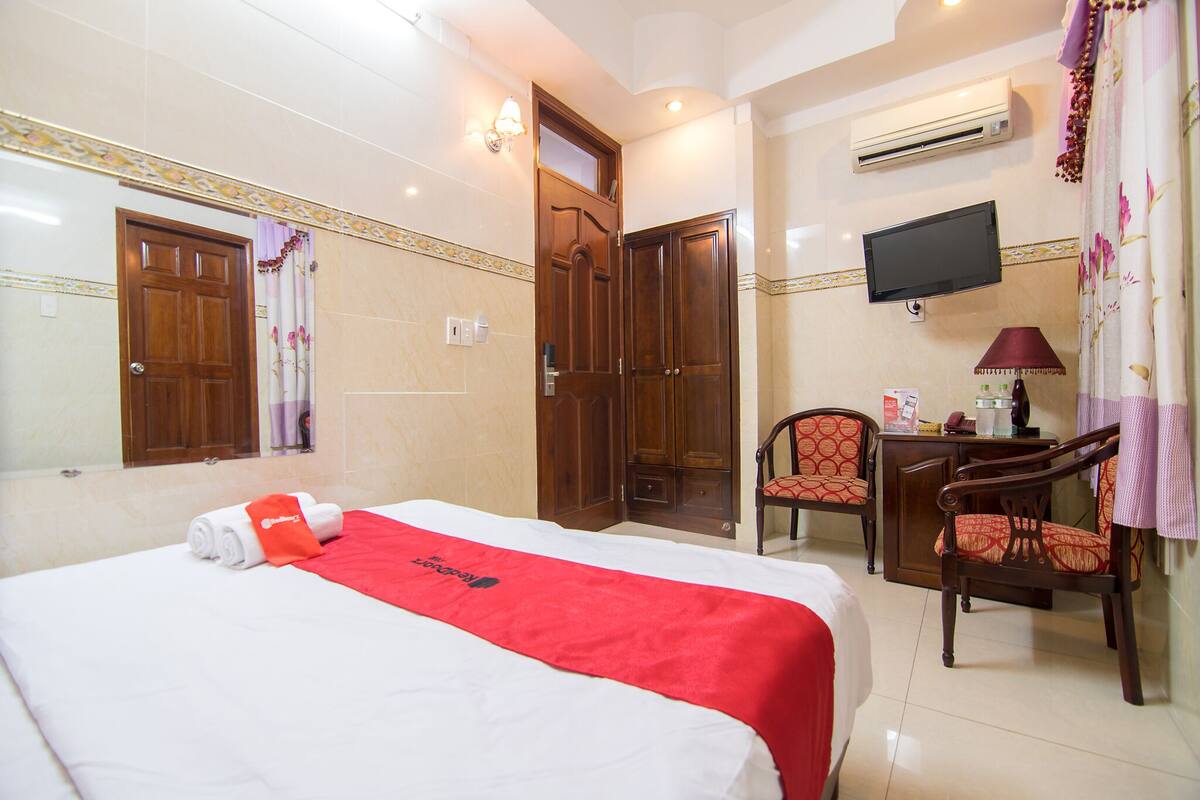 A hotel room with a bed, a desk, two chairs, a television, and an air conditioning unit.