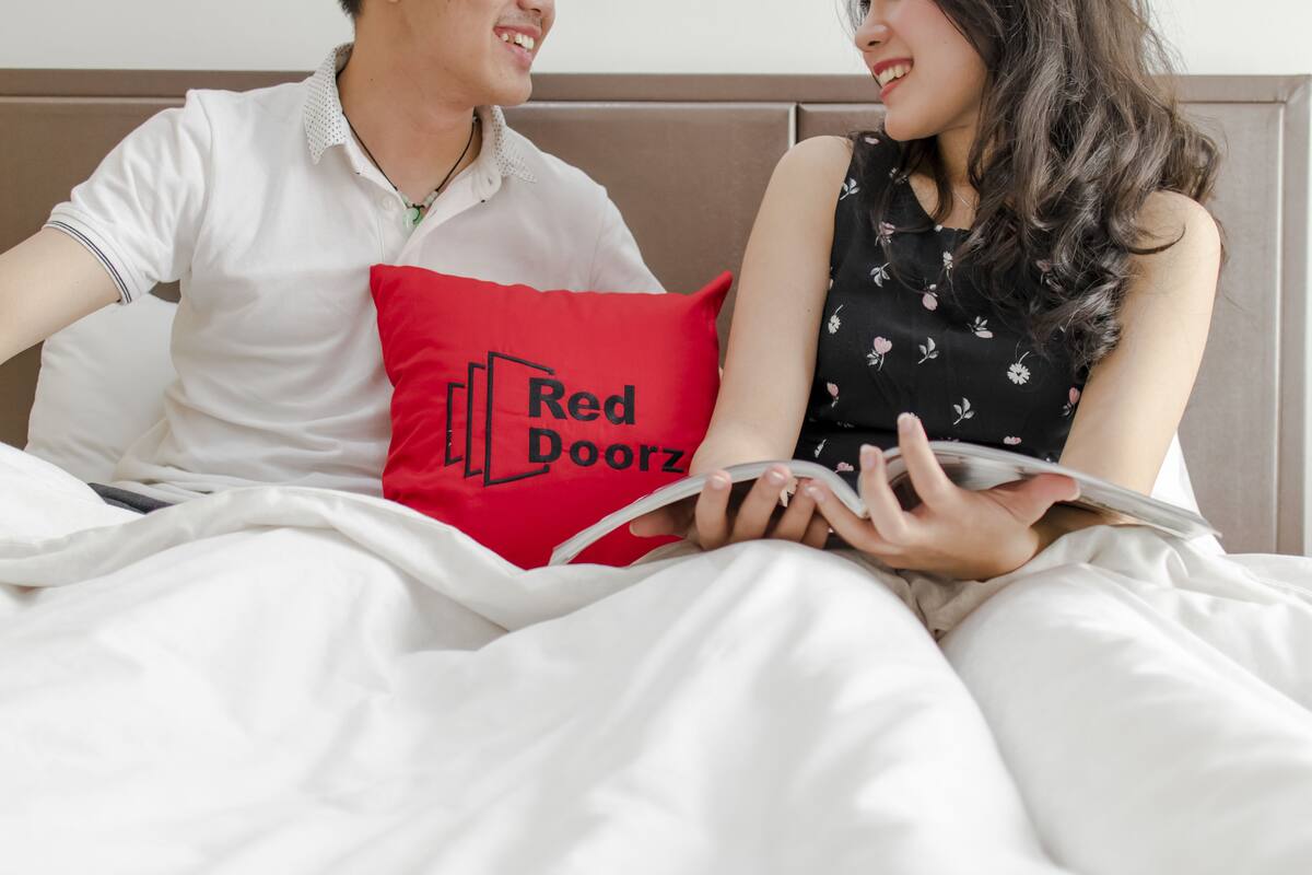 A couple sitting on a bed with a red pillow that has 'Red Doorz' printed on it.