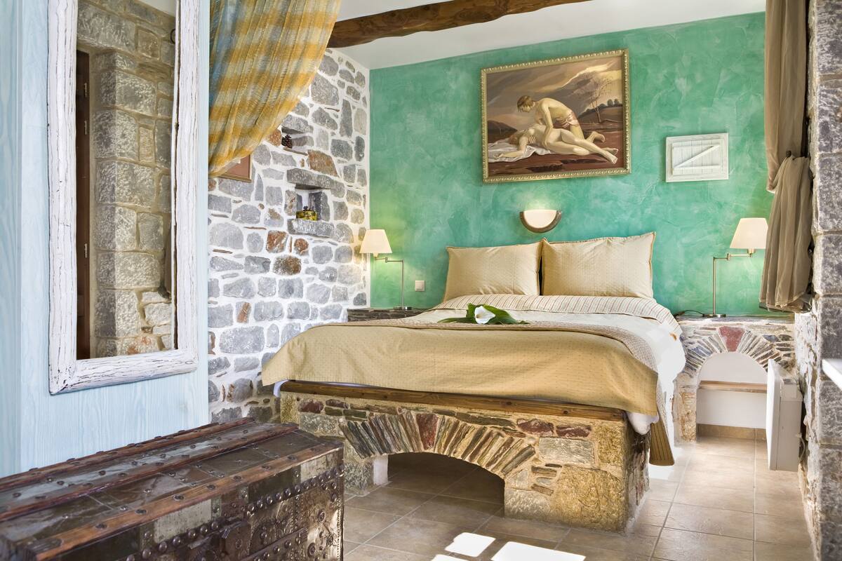 A bedroom with a stone wall, a bed with beige bedding, a painting above the bed, and a stone bench at the foot of the bed.
