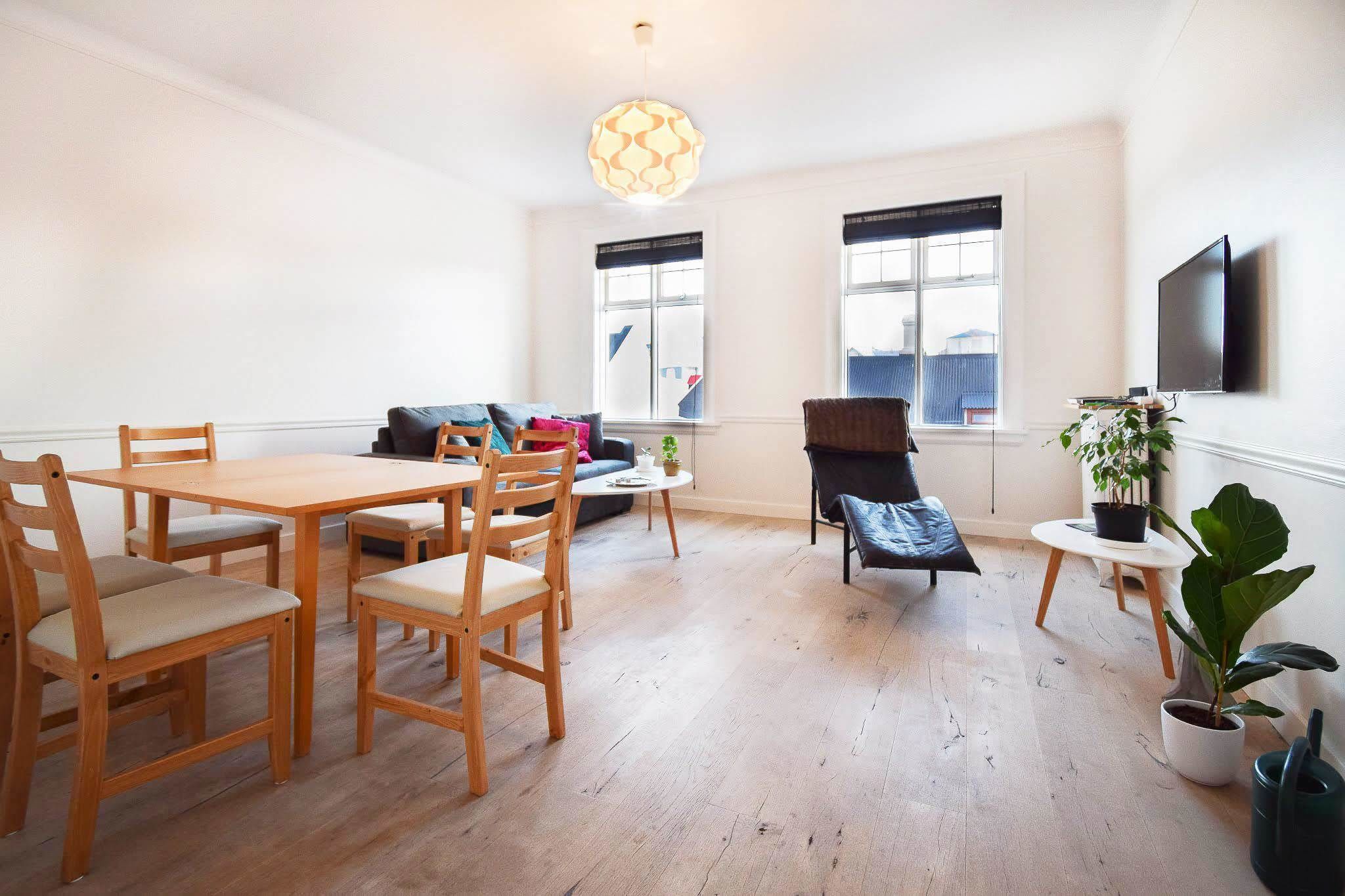 A spacious living room with a wooden dining table and chairs, a sofa, a TV mounted on the wall, and large windows.
