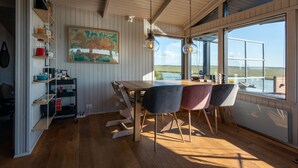 A dining area with a wooden table and chairs, a painting on the wall, and a view through large windows. - Paradise Cabin (Selfoss)