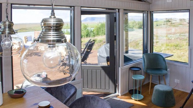 A cozy interior with a large glass lamp, a wooden table with cups, a blue chair, and a view of the outdoors through a door. - Paradise Cabin (Selfoss)