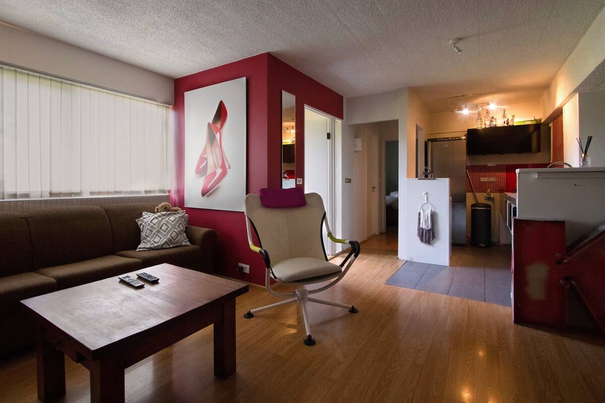 Ein modernes Wohnzimmer mit einer Couch, einem Holztisch und einem Sessel mit Kissen. Ein großes Gemälde hängt an der Wand, und im Hintergrund ist eine Küchenzeile zu sehen.