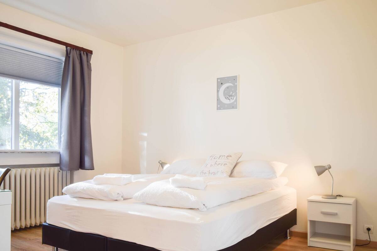 A neatly made bed with white linens and pillows, a bedside lamp, and a small bedside table.