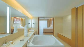 A modern hotel bathroom with a large bathtub, a vanity with a mirror, and a separate bedroom visible through an open door.