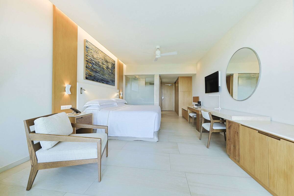 A modern hotel room with a bed, a desk, a chair, a large mirror, and a painting on the wall.