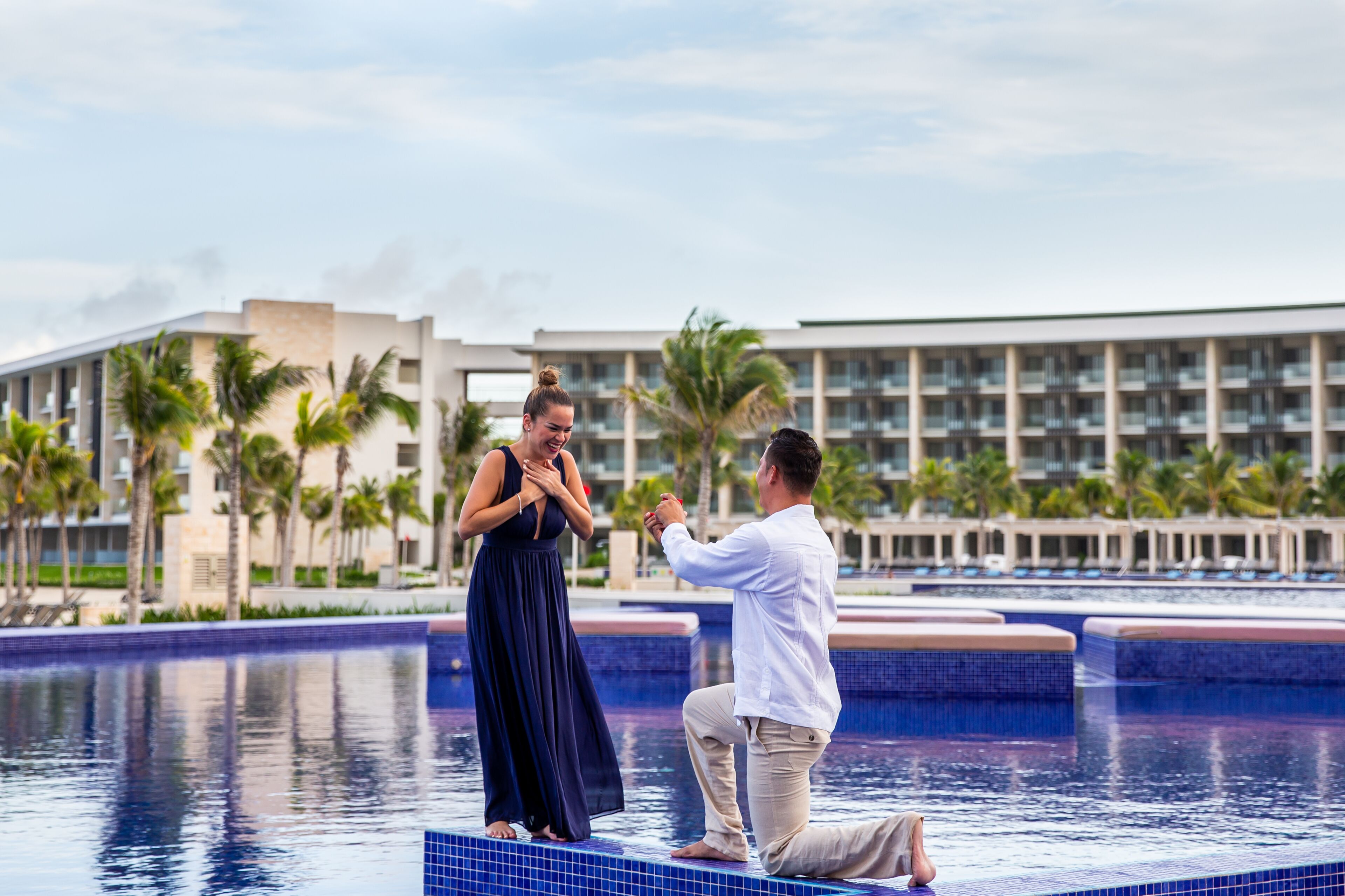 A man proposing to a woman by the poolside in a resort setting with palm trees and a modern building in the background.