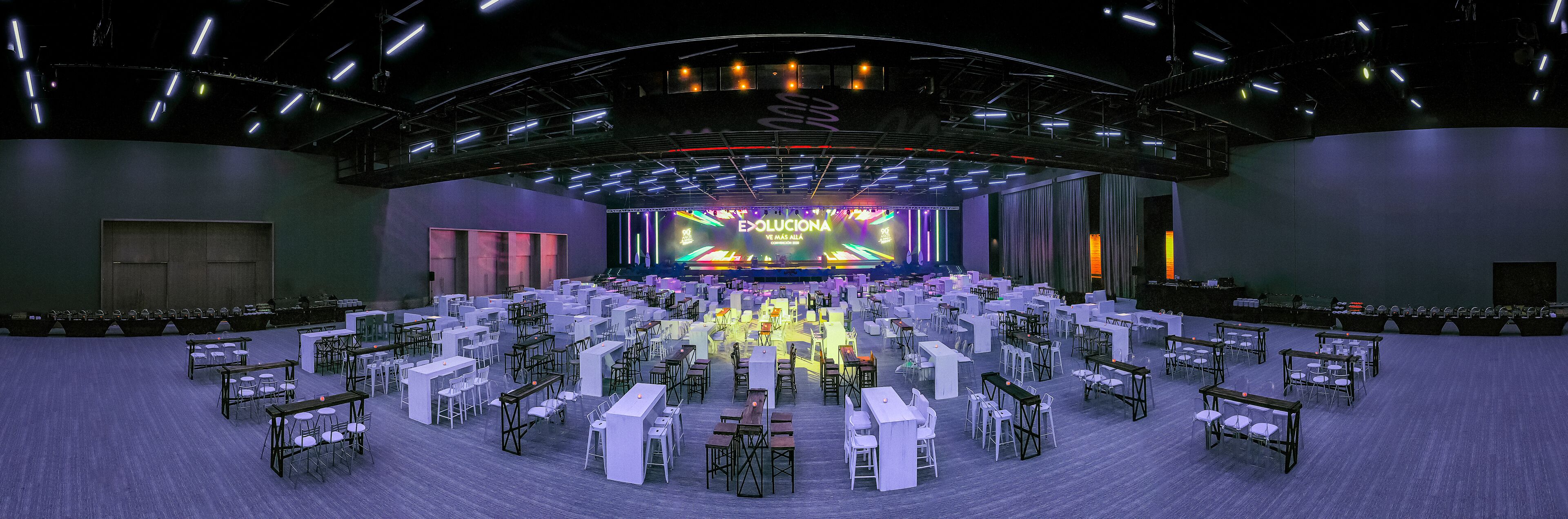 A large indoor venue with a stage, multiple tables and chairs arranged in rows, and a high ceiling with lighting fixtures.