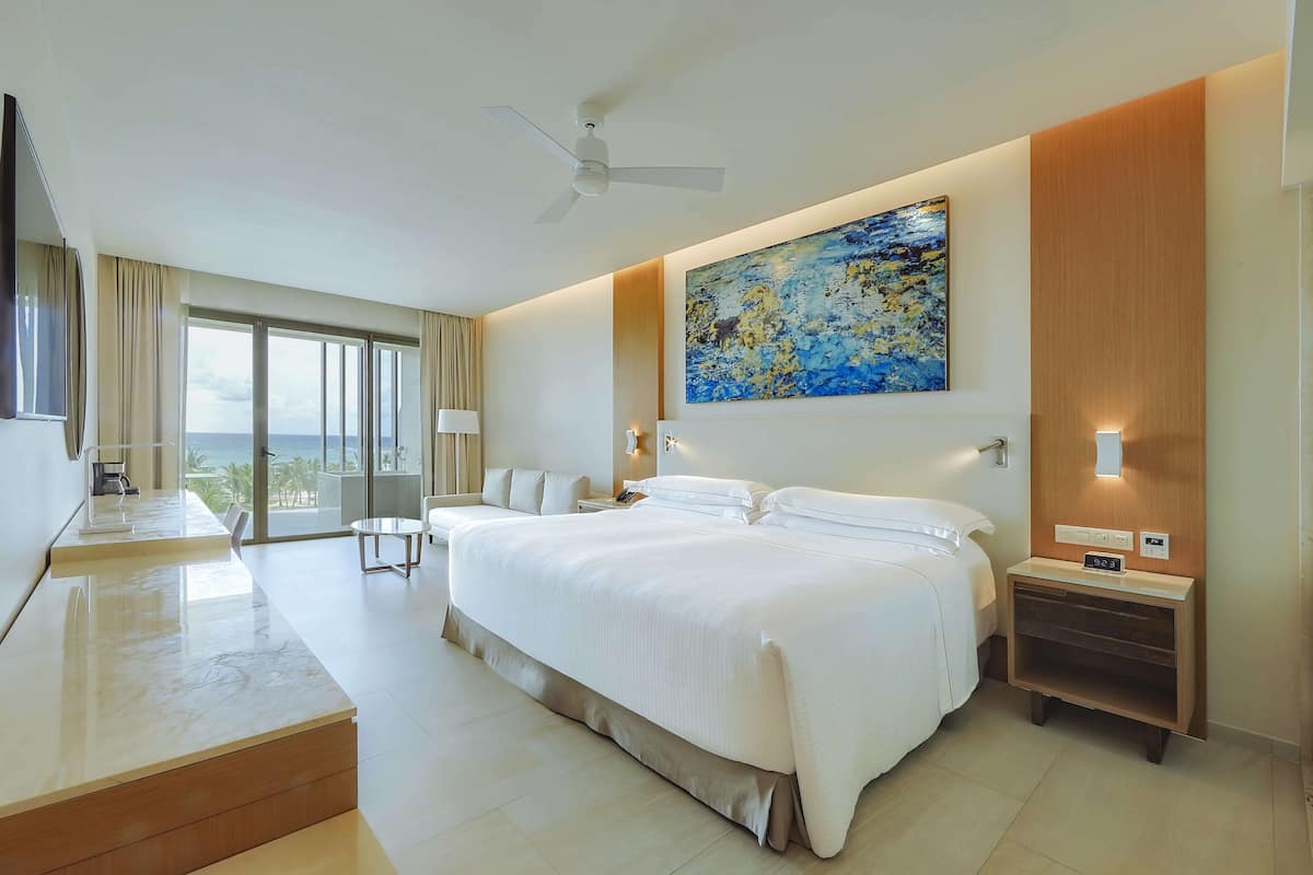 A modern hotel room with a large bed, a ceiling fan, a painting, and a view of the ocean.