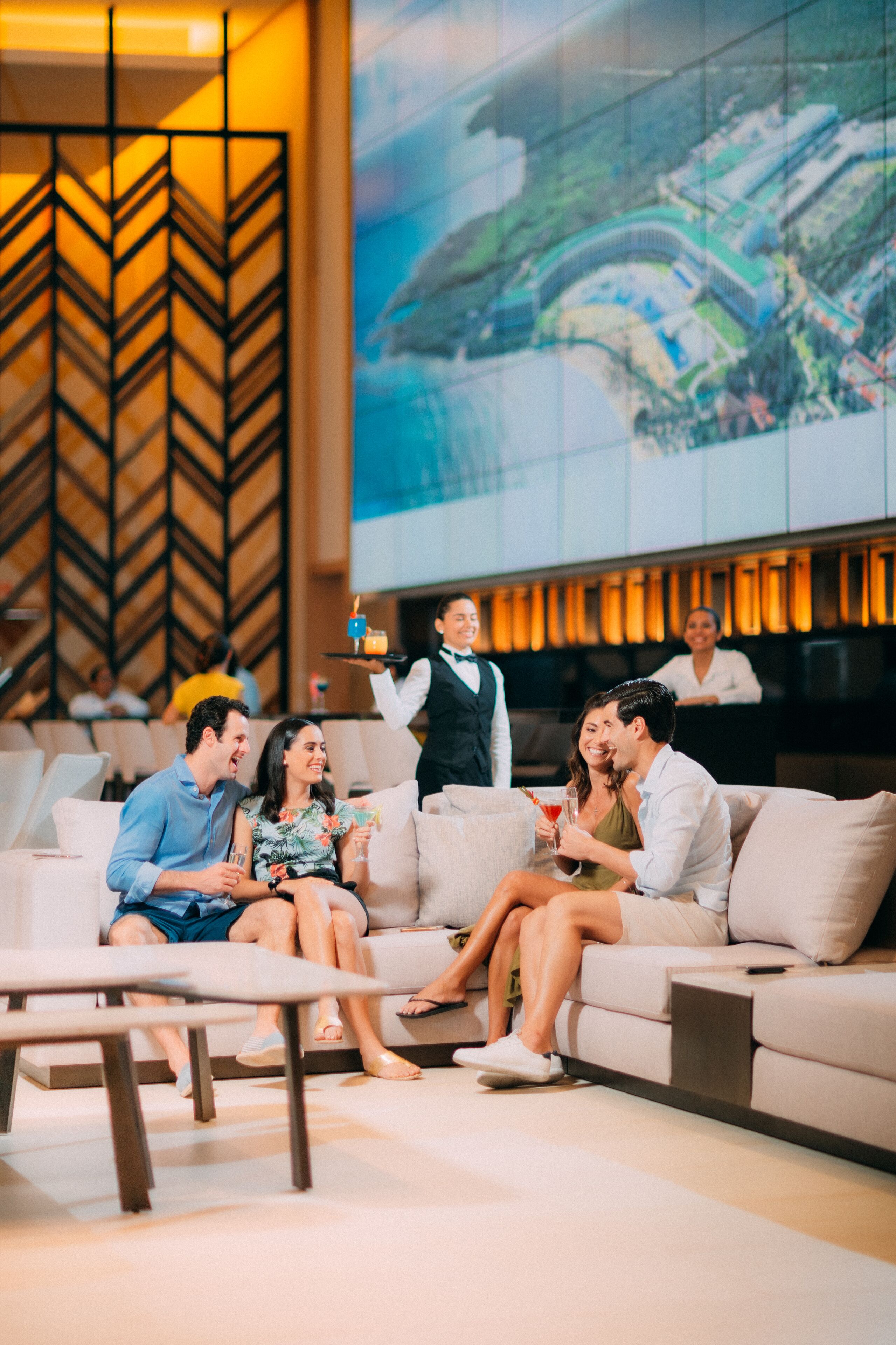 Un'area lounge moderna con un grande schermo che mostra un paesaggio, un divano con ospiti e un cameriere che serve bevande.