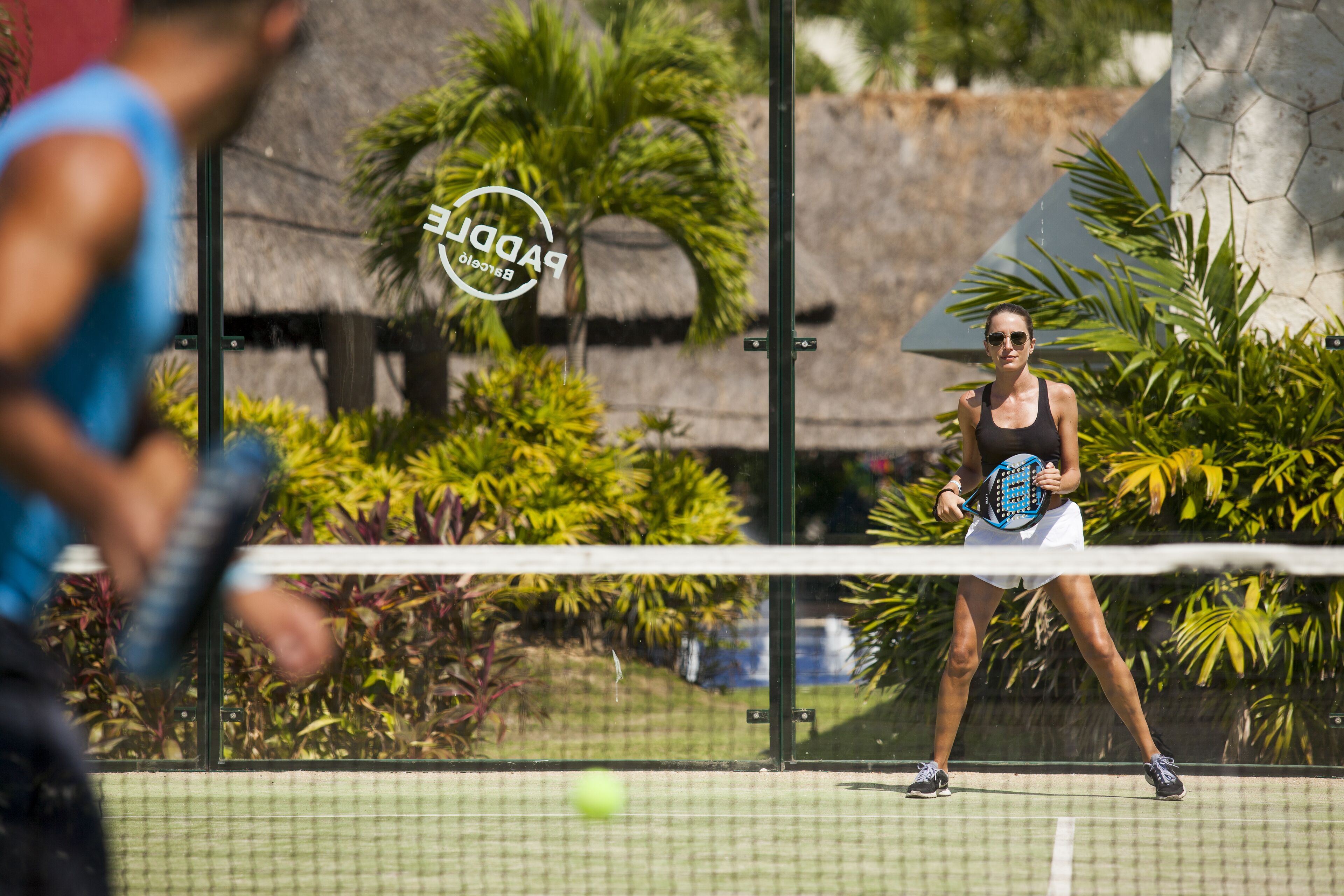 Una cancha de tenis con una red, palmeras y un edificio con techo de paja al fondo.