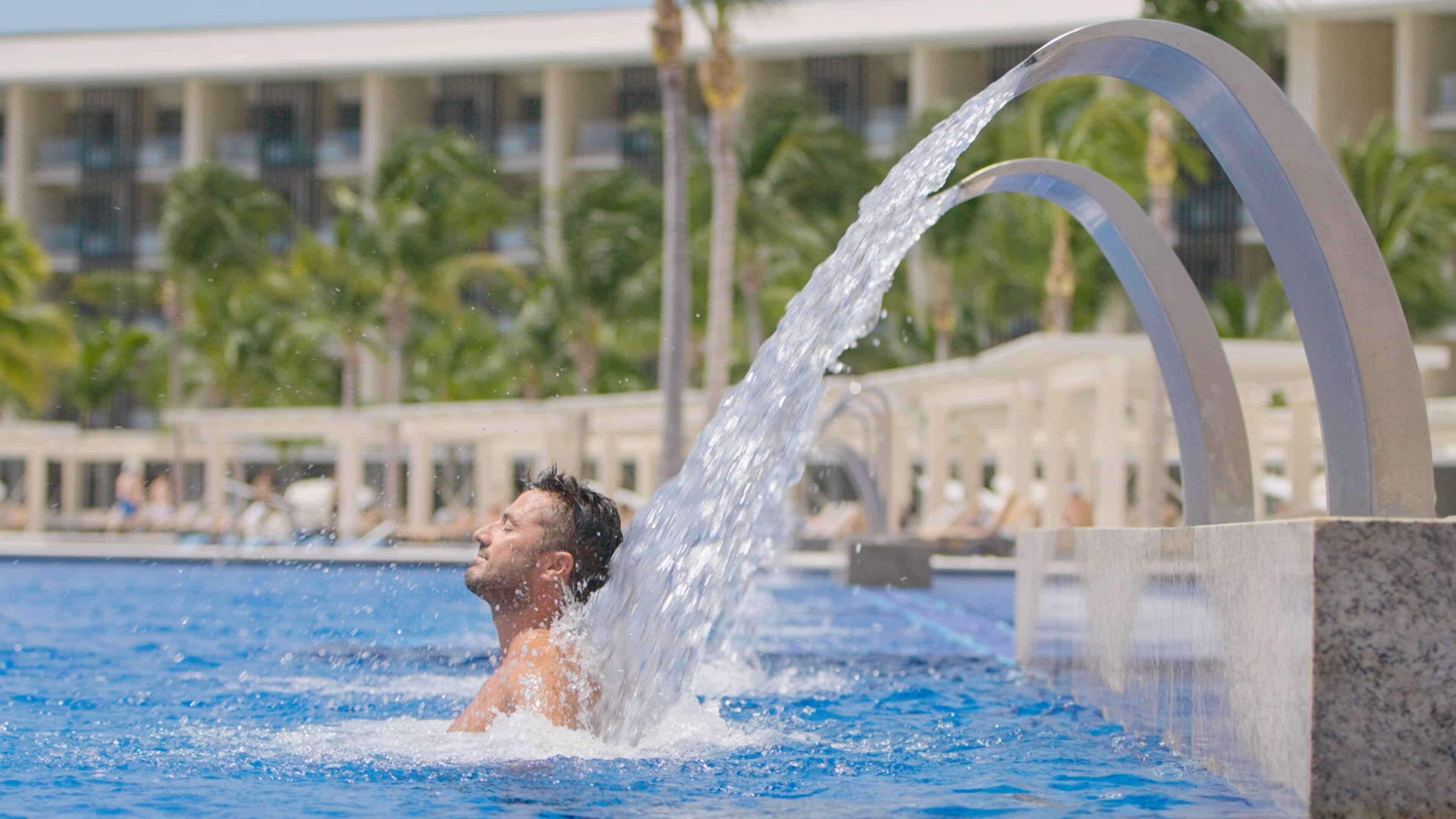 A man in a swimming pool with water flowing from a structure.