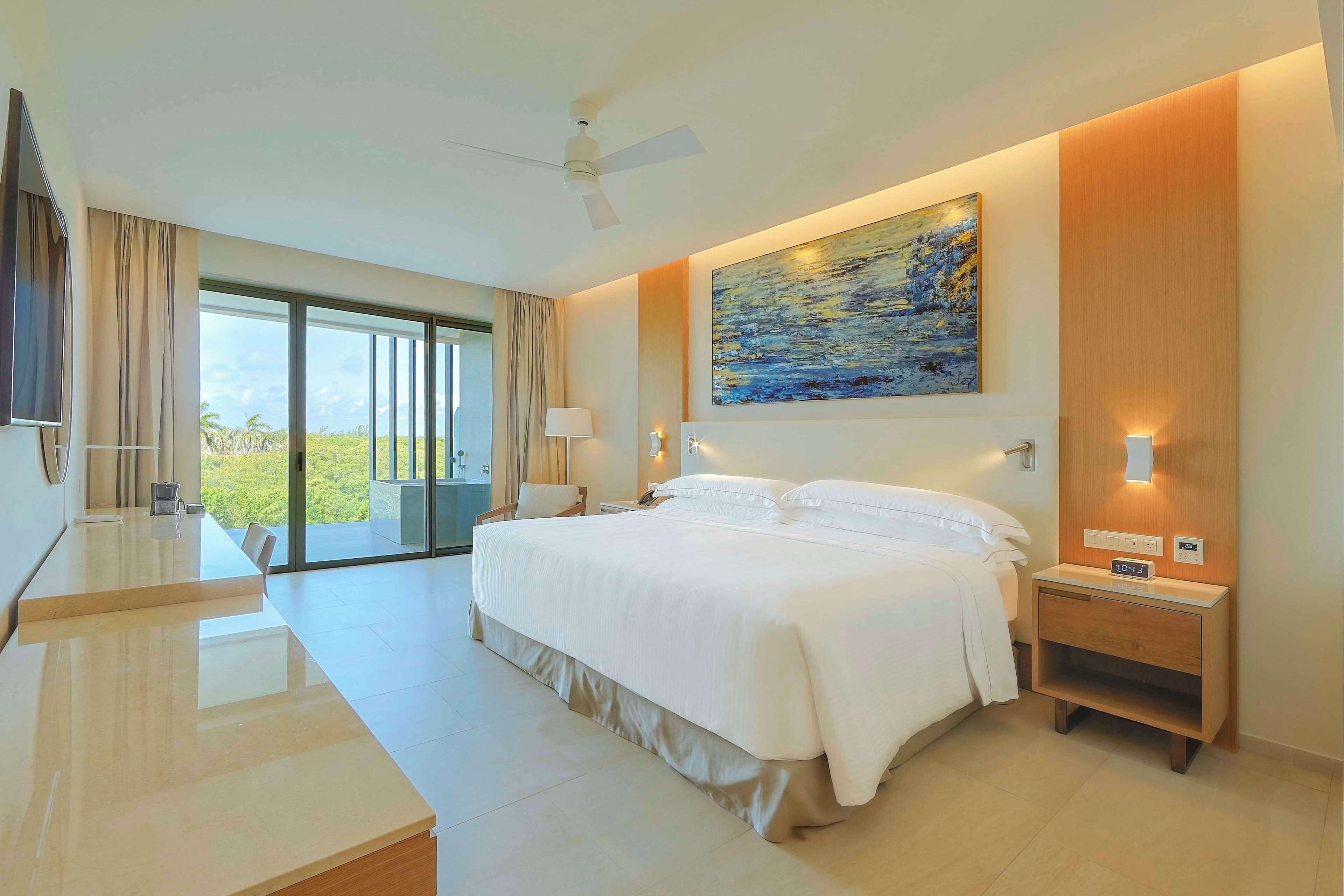A modern hotel room with a large bed, a desk, a chair, a TV, a painting, and a view of the outdoors.